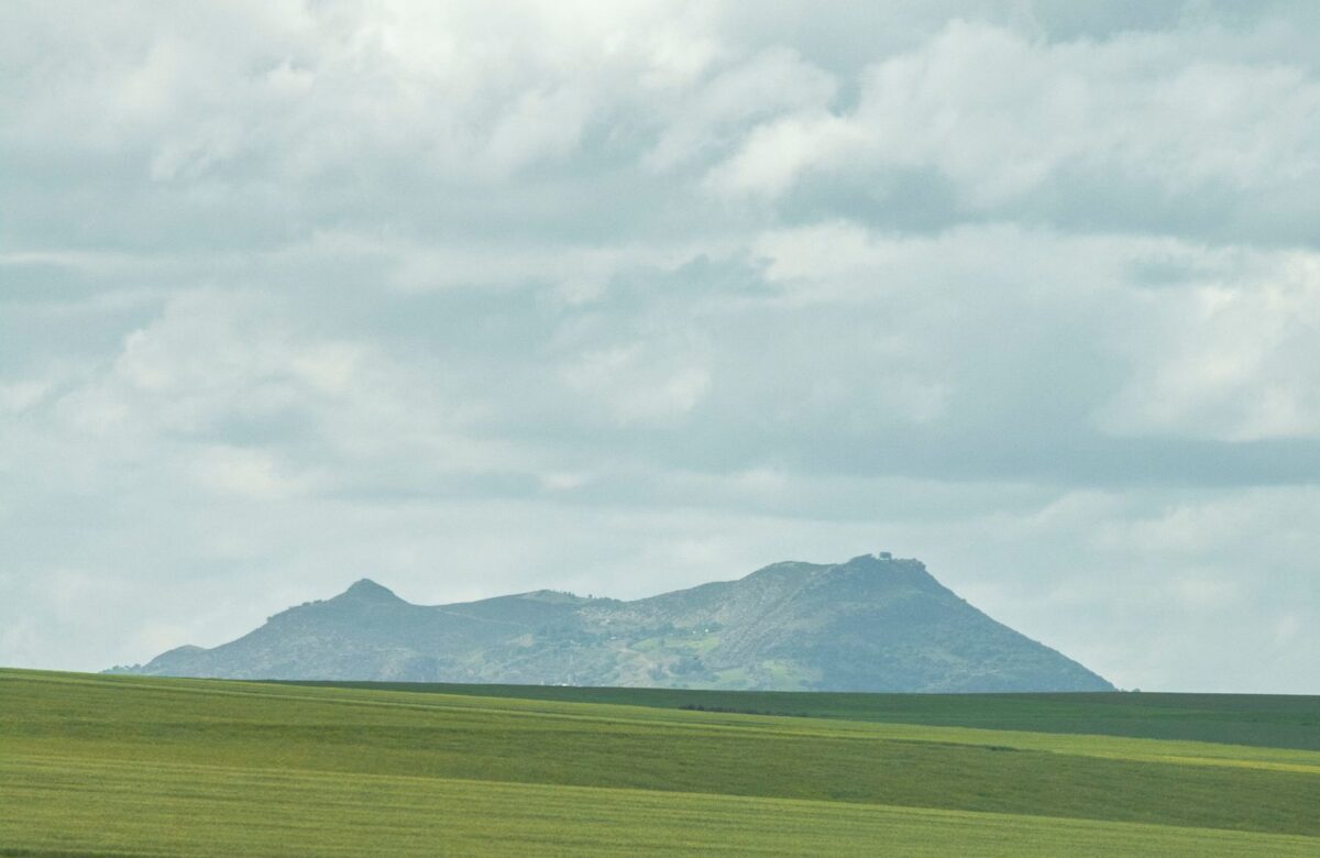 Green land and hills - LORDS OF THE LANDS Green open field beneath a distant forested hill under a cloudy sky, Sahyadri-style landscape — Lords of the Lands